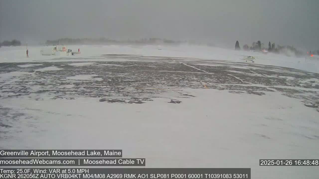 A snow-covered airfield is seen during a blizzard with a small plane visible in the distance.