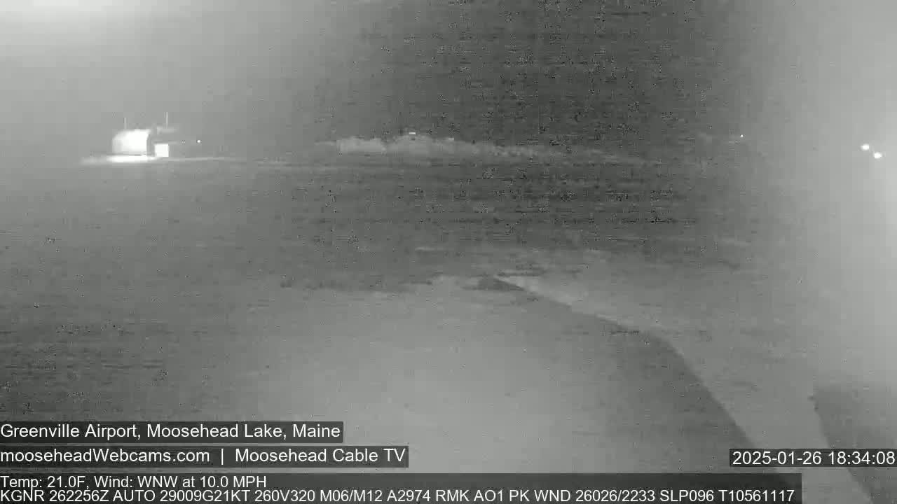 A blurry, nighttime grayscale image shows a sparsely lit airfield under a hazy sky with a wind of 10 mph from the west-northwest.