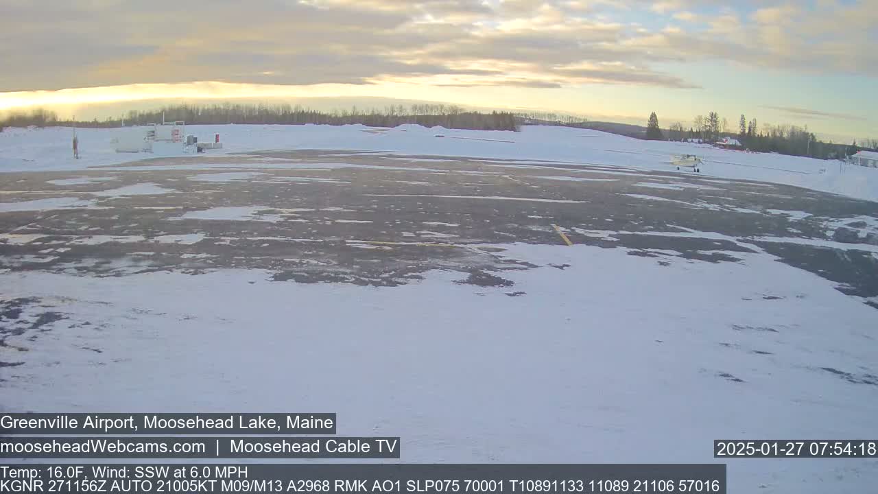 A small airplane sits on a snow-covered tarmac at an airport under a partly cloudy sky.