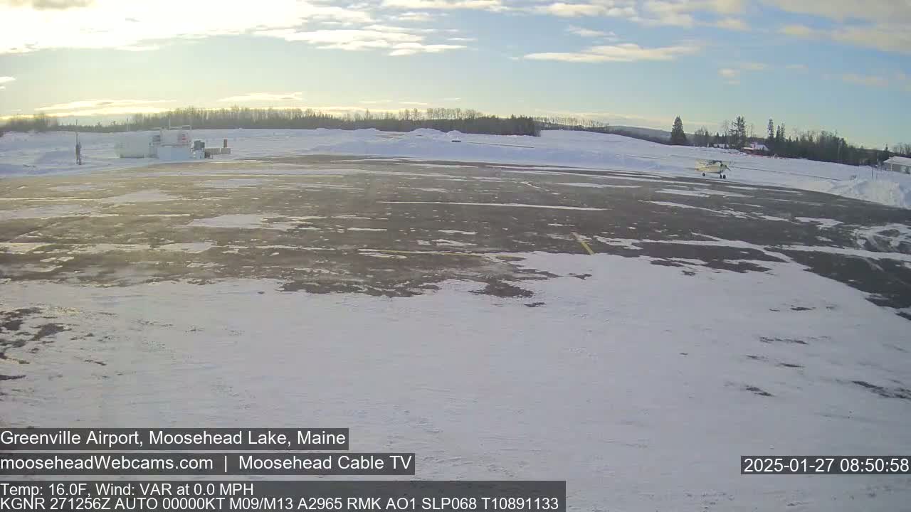 A small airplane sits on a snow-covered tarmac at a small airport, under a mostly clear sky.