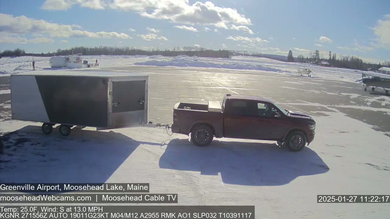 A maroon pickup truck towing a gray and white enclosed trailer is parked on a snow-covered airfield under a mostly sunny sky.