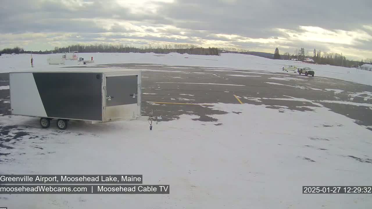 A gray and white enclosed trailer sits in a snow-covered airport parking lot under a partly cloudy sky.