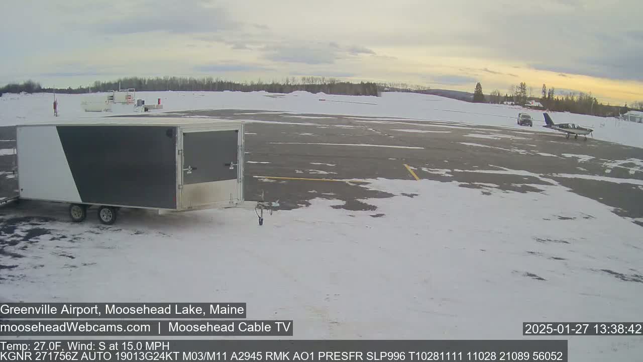 A gray and white enclosed trailer sits on a snowy airport tarmac next to a small airplane under a partly cloudy sky.