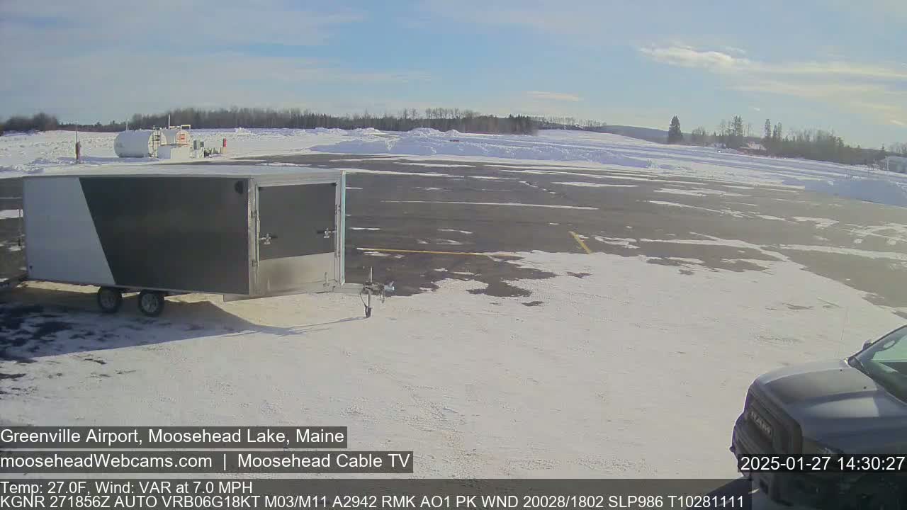 A gray enclosed trailer and part of a dark-colored truck are parked on a snow-covered tarmac in front of a distant line of trees under a mostly sunny sky.