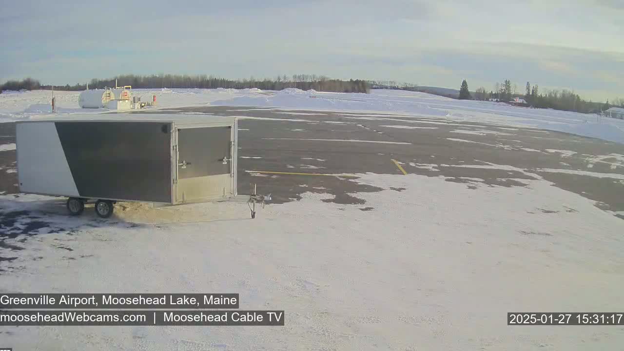 A gray and white enclosed trailer sits in a snowy airport parking lot under a partly cloudy sky.