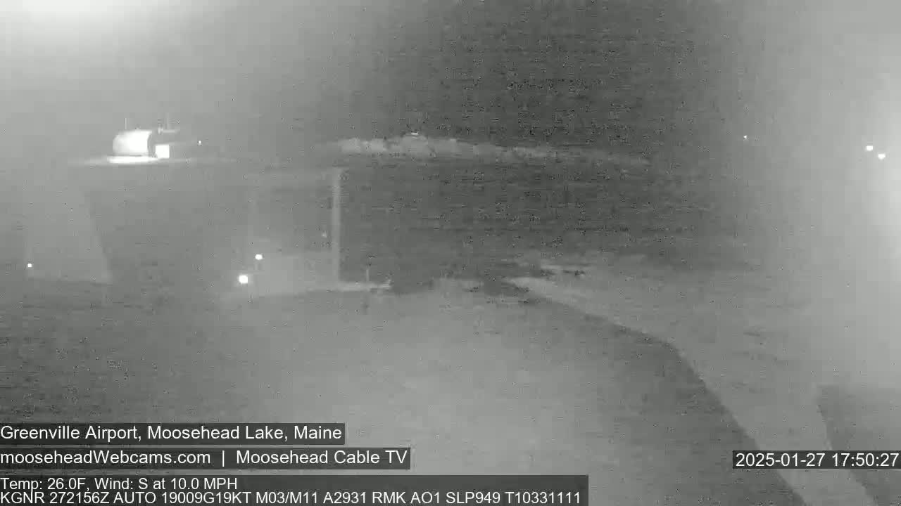 A nighttime, low-visibility view of a mostly empty airfield with a building in the background, under a hazy sky with a south wind of 10 mph and a temperature of 26 degrees Fahrenheit.