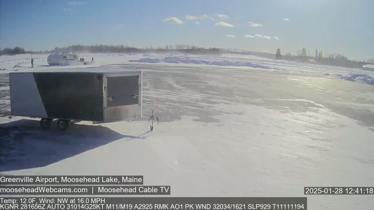 A gray and white enclosed trailer sits in a snow-covered airfield under a mostly sunny sky, with a light wind blowing.