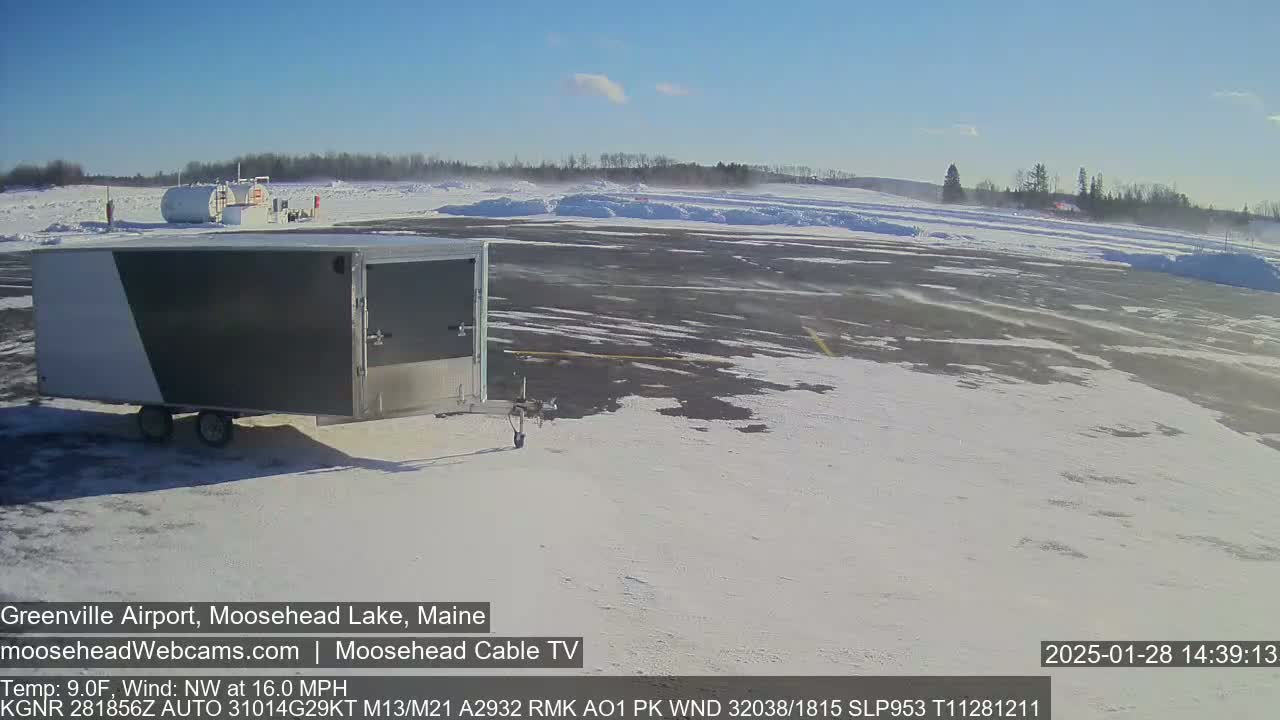 A gray and white enclosed trailer sits in a snow-covered airport tarmac on a sunny day with a northwest wind of 16 mph.