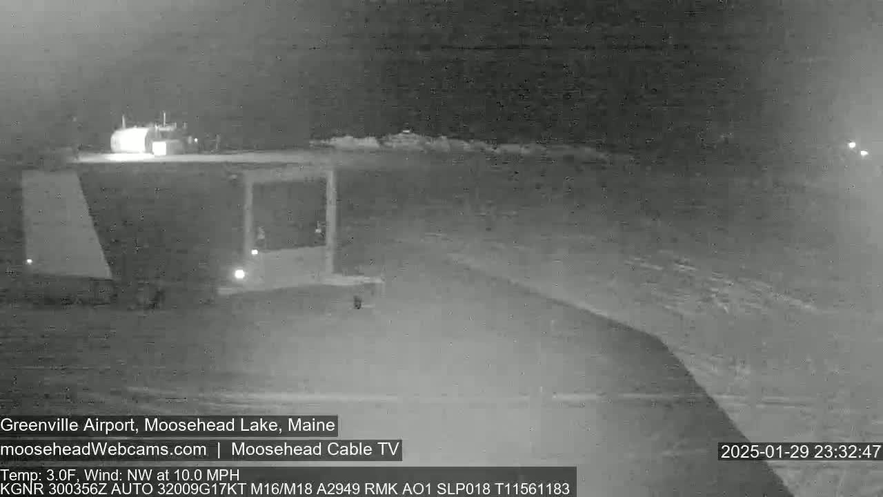 A nighttime, low-light image shows a snow-covered airport with a building and a few lights, under a dark, possibly cloudy sky with a northwest wind.