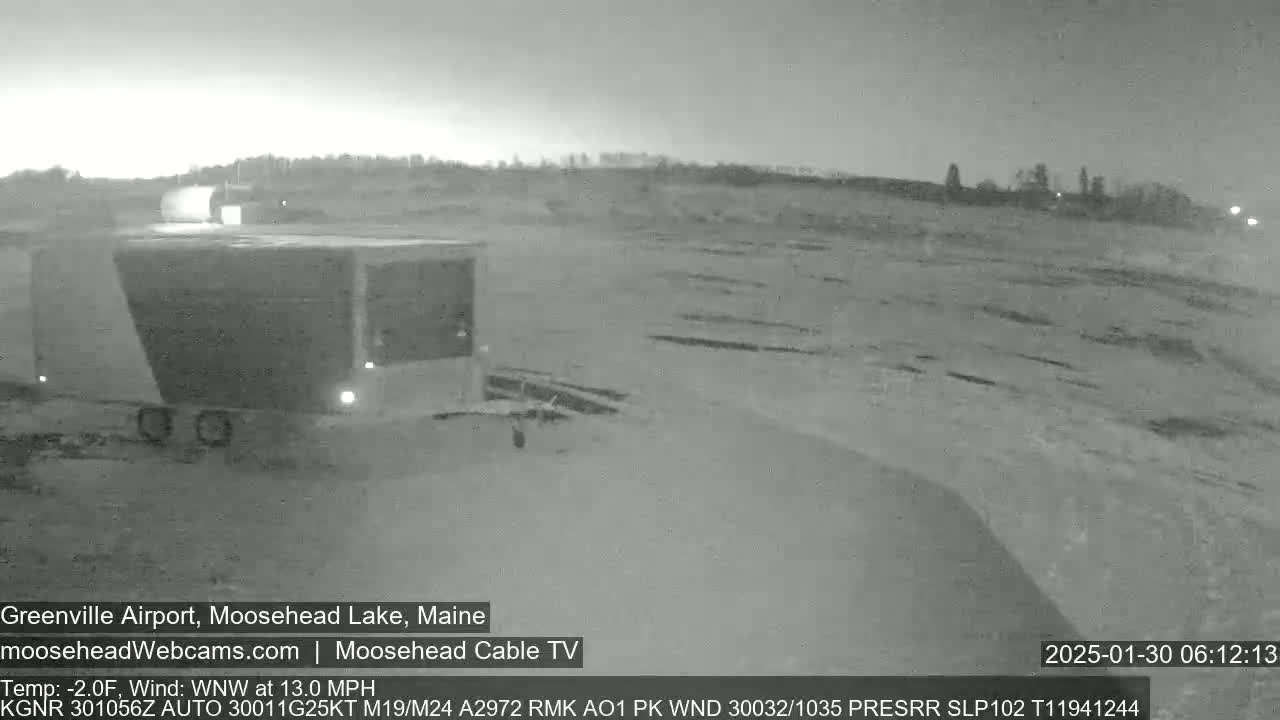 A nighttime, low-light grayscale image shows a mostly empty airfield with some buildings and vehicles under a dark sky with a northwest wind at 13 mph and a temperature of -2°F.