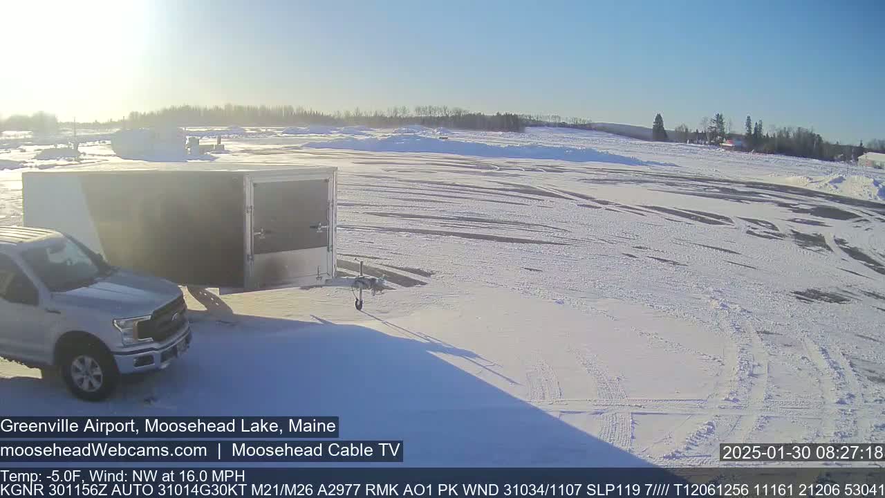A silver pickup truck is parked next to a large white enclosed trailer in a snow-covered airport parking lot under a clear, sunny sky.