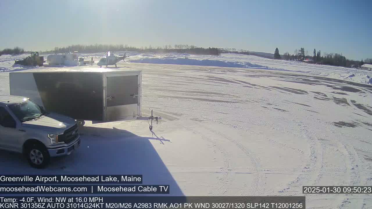 A helicopter sits on a snow-covered airfield next to a pickup truck towing a large cargo trailer, under a bright, sunny sky.