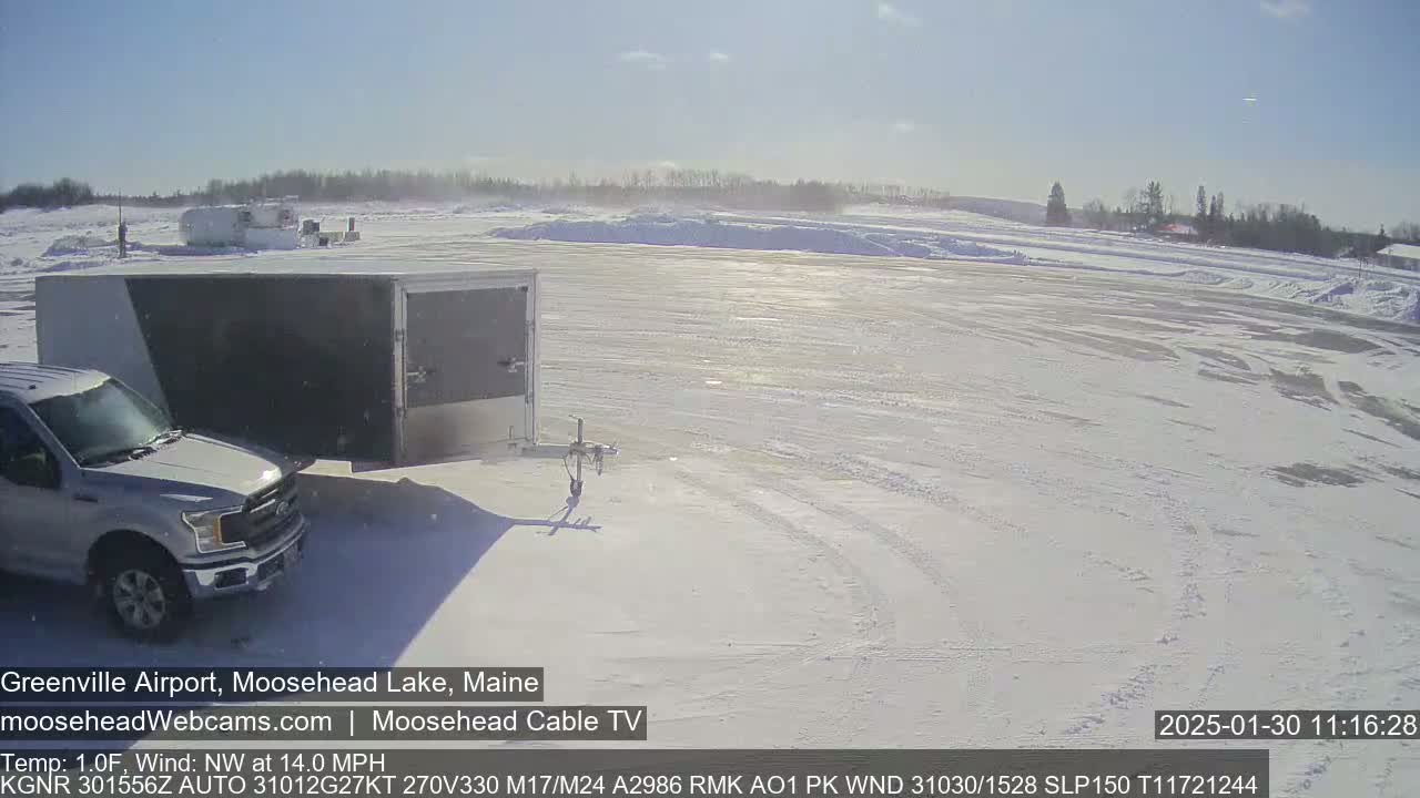 A white pickup truck towing a gray enclosed trailer is parked on a snow-covered airfield under a bright, sunny sky with a northwest wind.