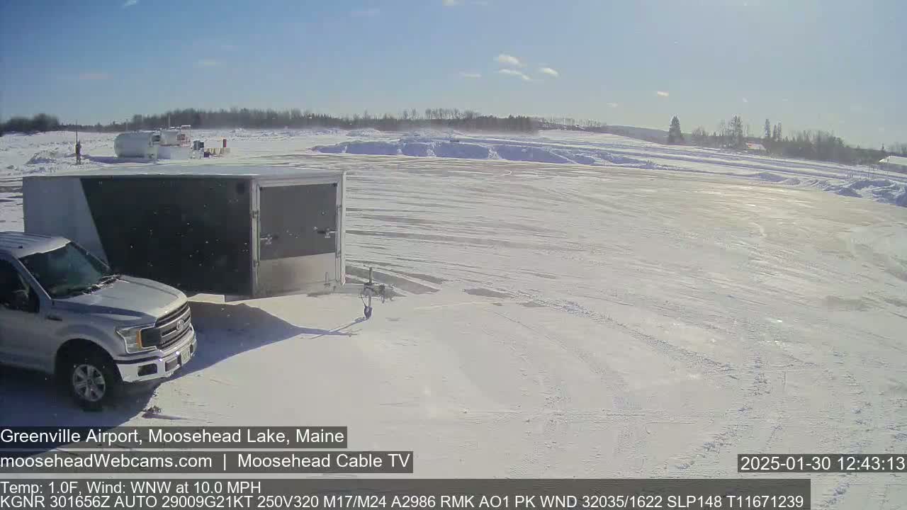 A silver pickup truck towing a white enclosed trailer is parked in a snow-covered airport lot under a clear blue sky.