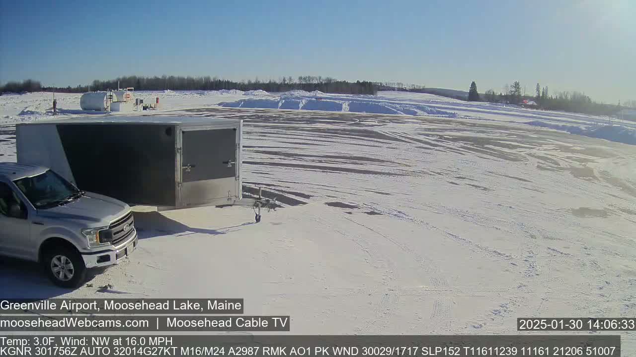 A silver pickup truck is parked next to a gray enclosed trailer in a snow-covered airport parking lot under a clear, sunny sky.