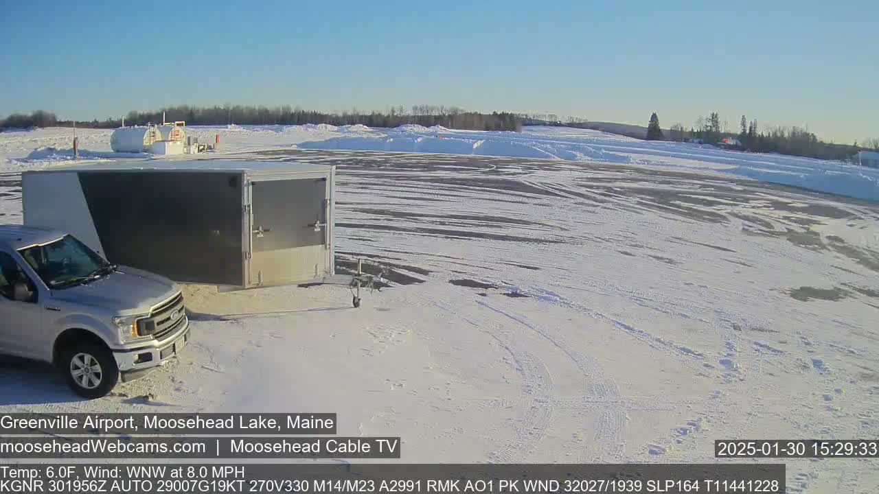 A silver pickup truck is parked next to a large, enclosed trailer in a snow-covered airport parking lot under a clear, sunny sky.