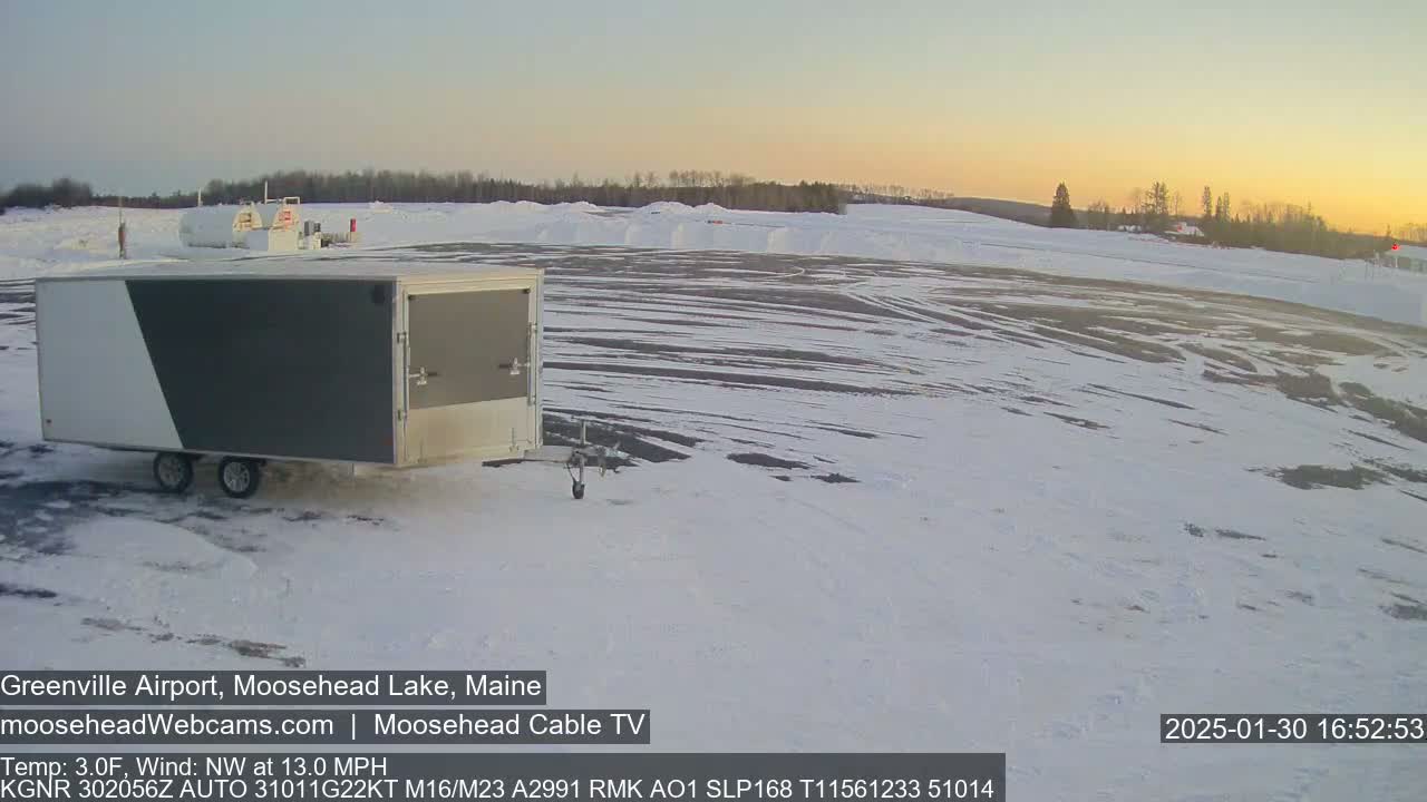 A gray and white enclosed trailer sits in a snow-covered airport parking lot during a sunset with a northwest wind at 13 mph and a temperature of 3 degrees Fahrenheit.