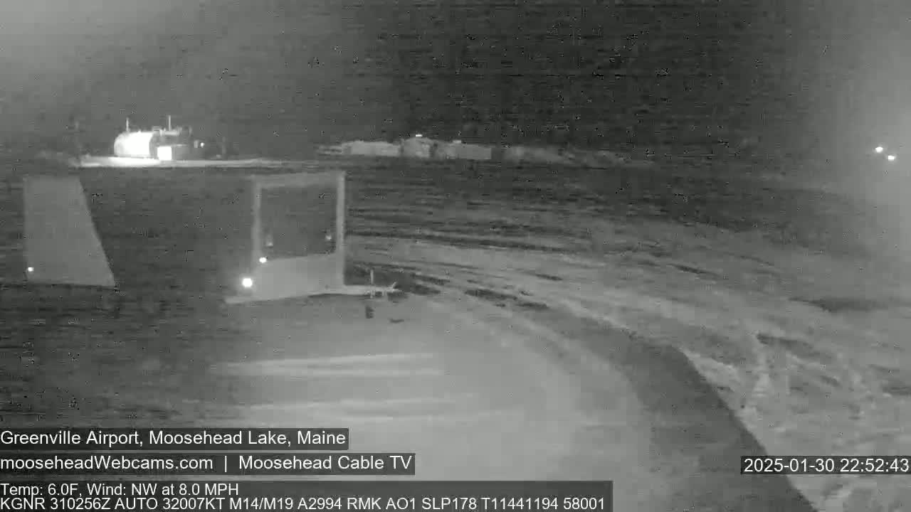 A nighttime, snowy scene shows a mostly empty airport runway with a few vehicles visible under a dark sky.