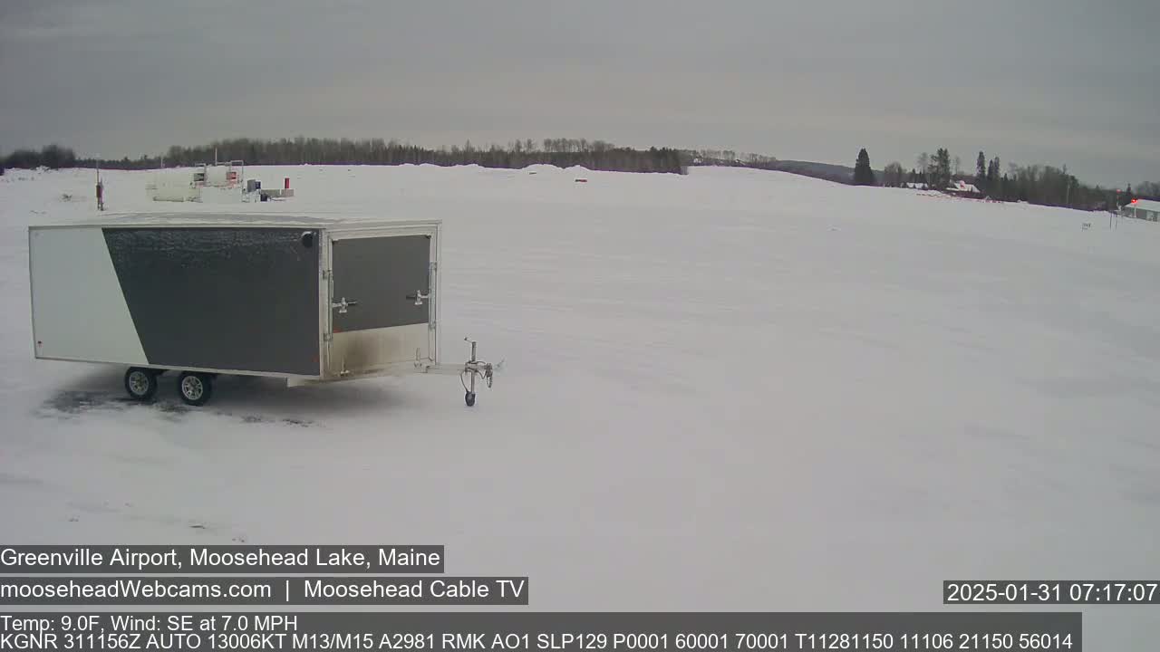 A gray and white enclosed trailer sits in a snow-covered airfield under an overcast sky.