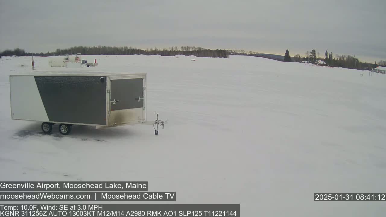 A gray and white enclosed trailer sits in a snow-covered airfield under a cloudy sky.