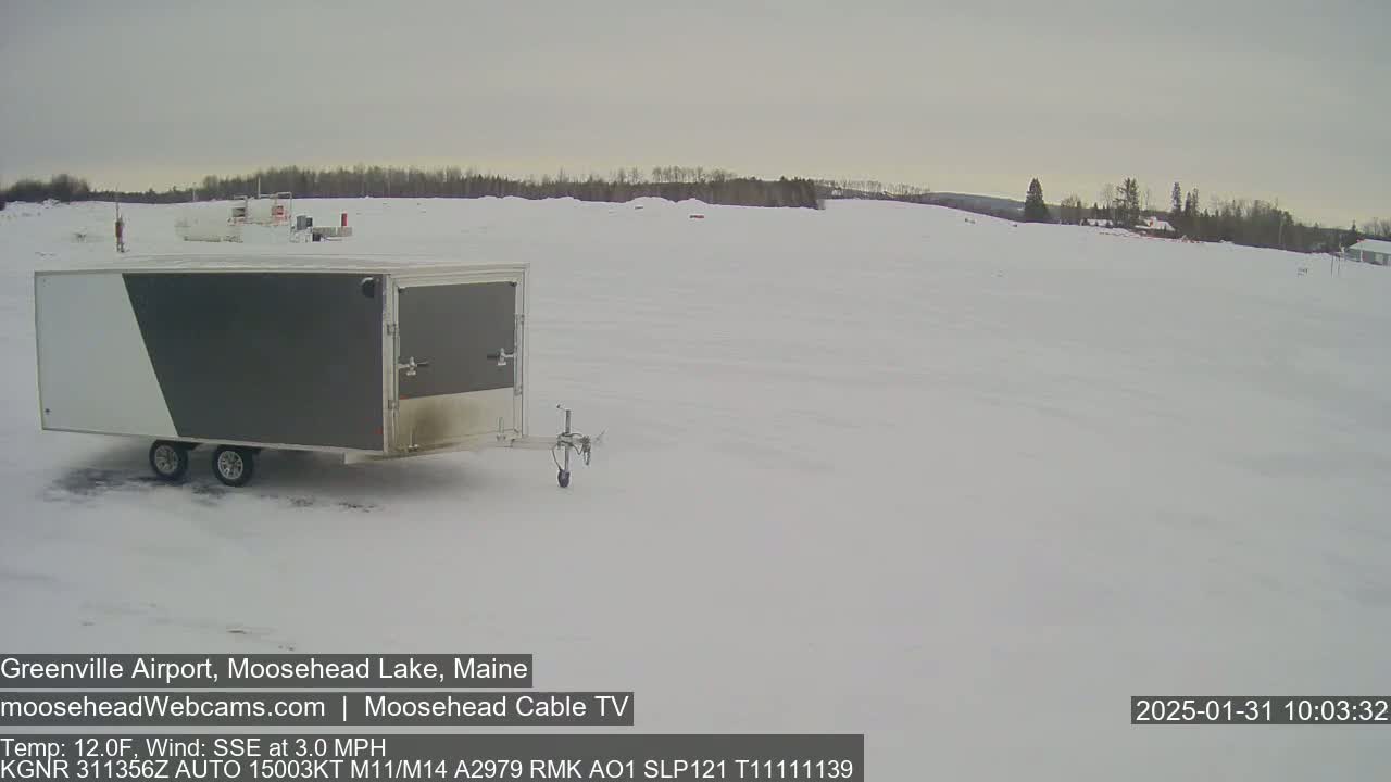 A gray and white enclosed trailer sits in a snow-covered field under an overcast sky.