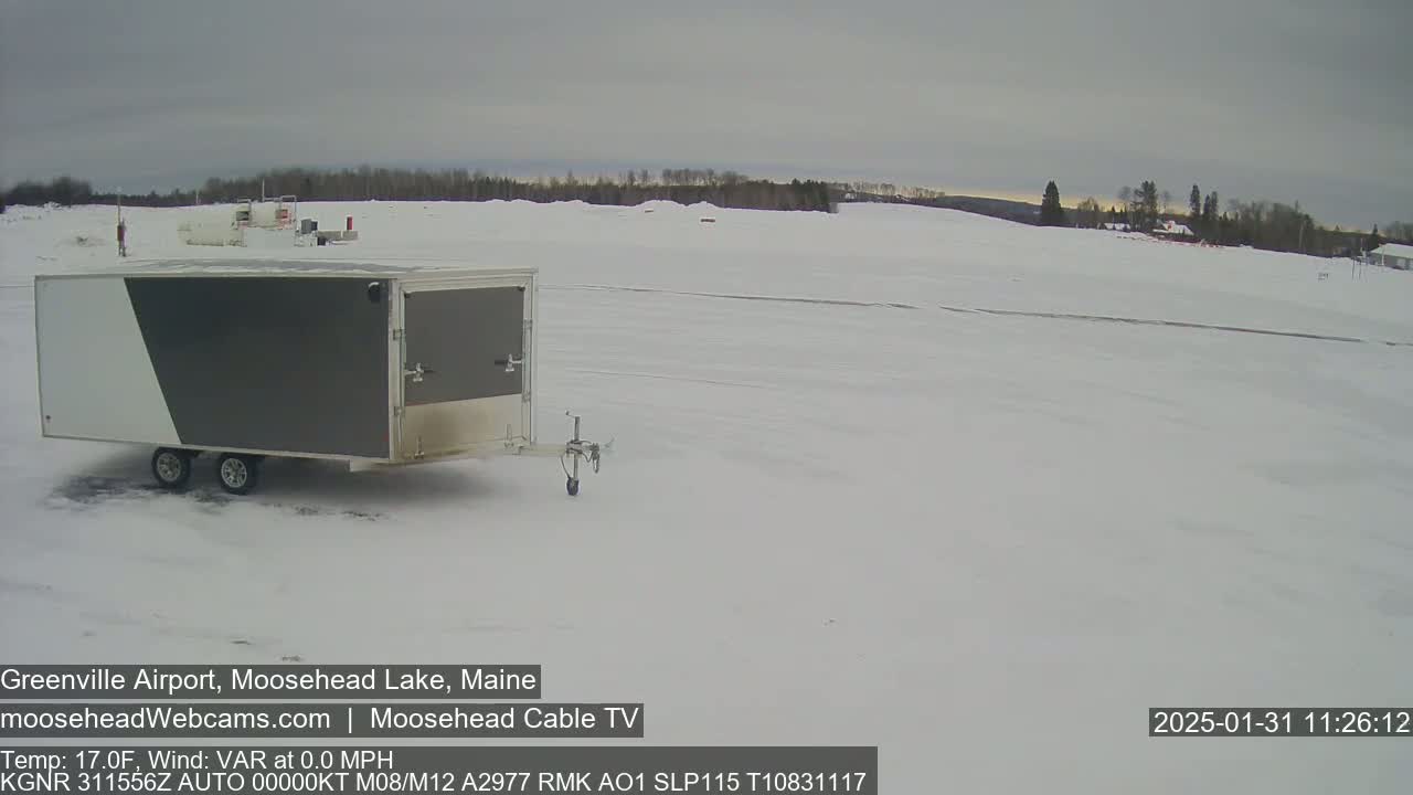 A gray and white enclosed trailer sits in a snow-covered airfield under an overcast sky.