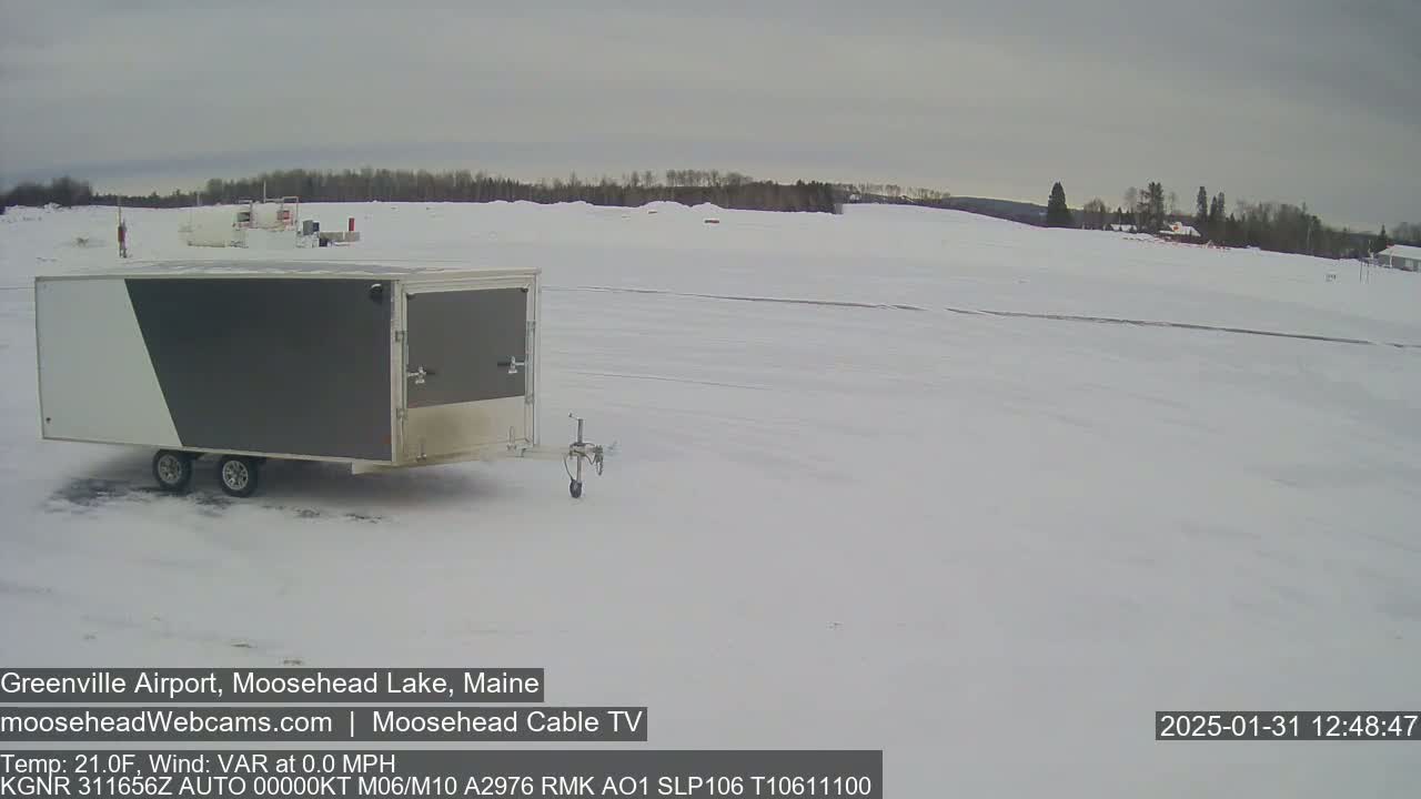 A gray and white enclosed trailer sits in a snow-covered airfield under an overcast sky.