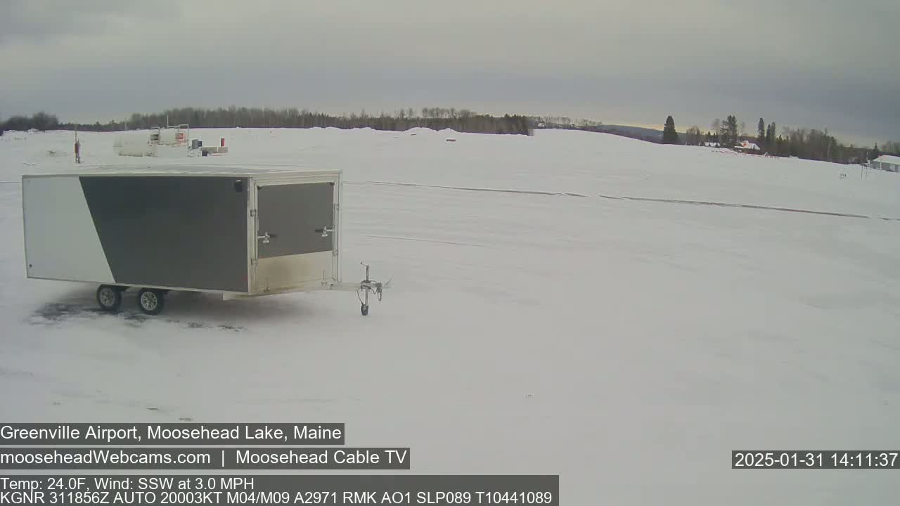 A gray and white enclosed trailer sits in a snow-covered airfield under an overcast sky.