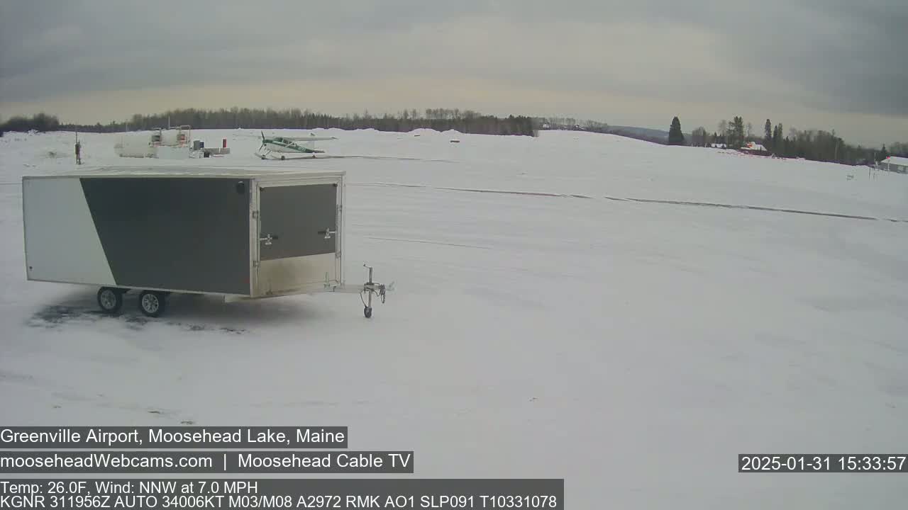 A gray and white enclosed trailer sits in a snow-covered airfield near a small airplane under an overcast sky.