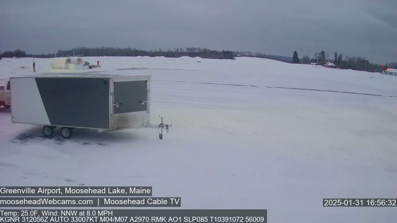A gray and white enclosed trailer is parked in a snow-covered airfield under a cloudy sky.