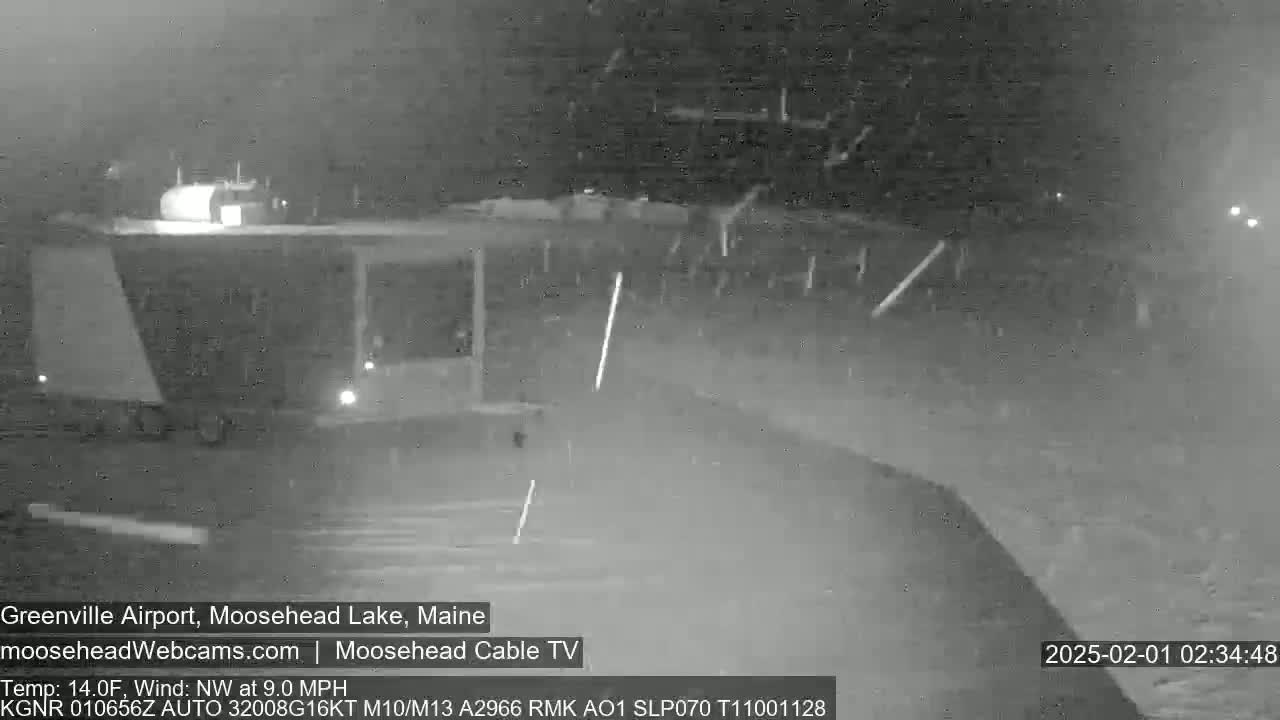 A nighttime, snowy scene shows a mostly empty parking lot with a large truck parked near a building.