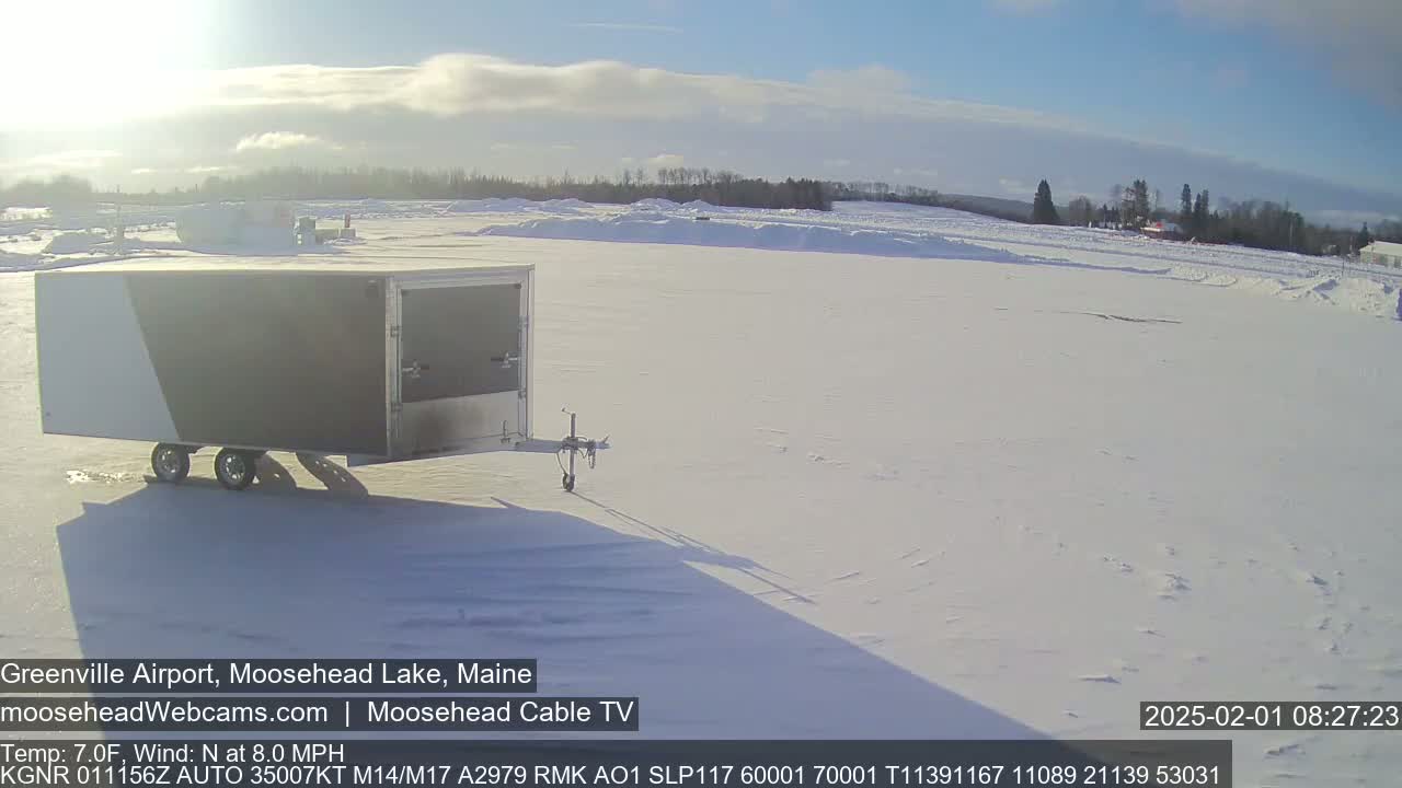 A gray and white enclosed trailer sits on a snow-covered airfield under a mostly sunny sky.