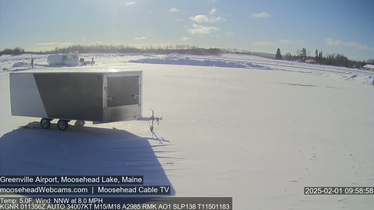 A gray and white enclosed trailer sits on a snow-covered expanse under a bright, sunny sky.