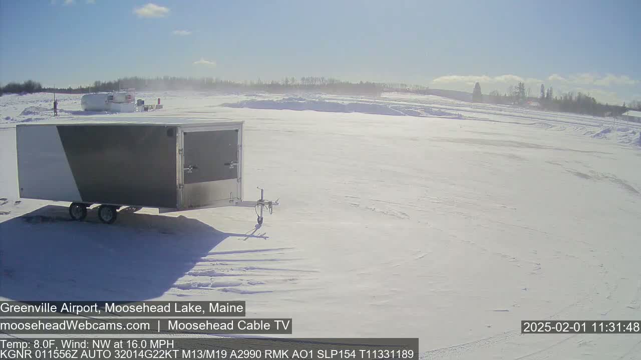 A gray and white enclosed trailer sits in a snow-covered field under a bright, sunny sky with a northwest wind.