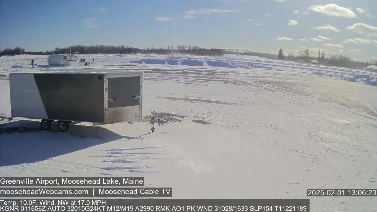 A gray and white enclosed trailer sits in a snowy airfield under a sunny sky.