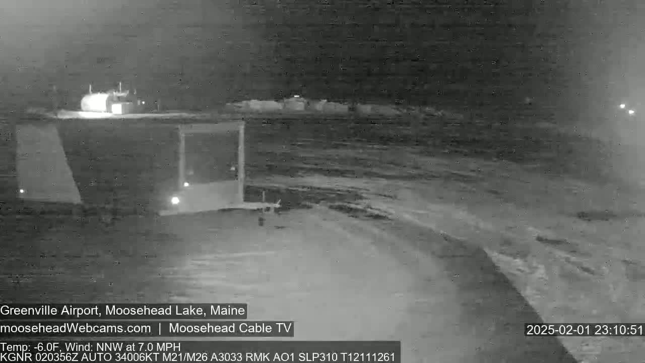A nighttime, snow-covered scene shows a building and a portion of a road or runway with a 7 mph wind from the northwest at -6°F.