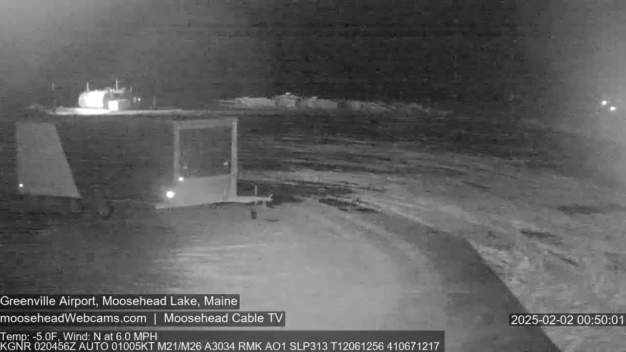 A nighttime, snow-covered parking lot is shown with a small building and a trailer visible under a dark sky.