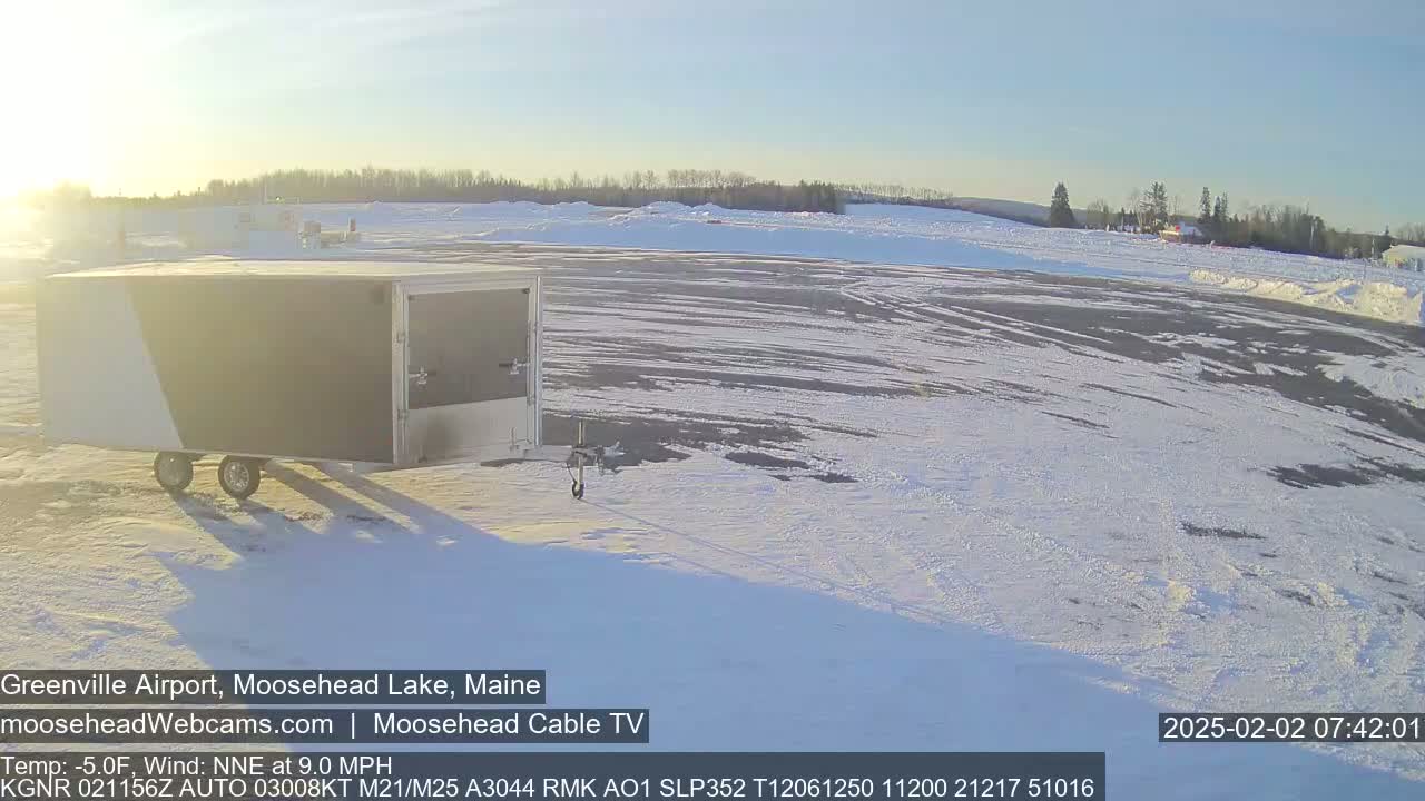A white and gray enclosed trailer sits in a snow-covered airport parking lot on a sunny, cold morning.