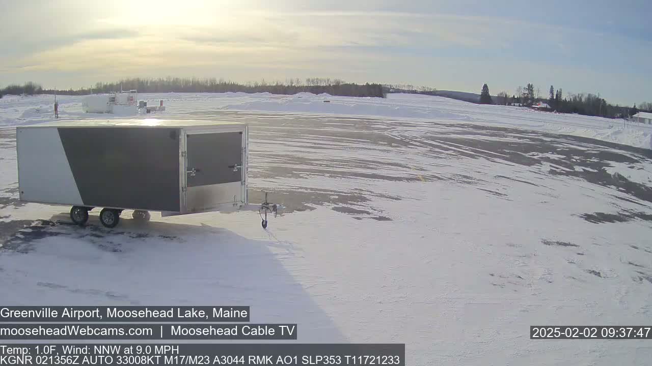 A gray and white enclosed trailer sits in a snowy airport parking lot under a partly sunny sky.