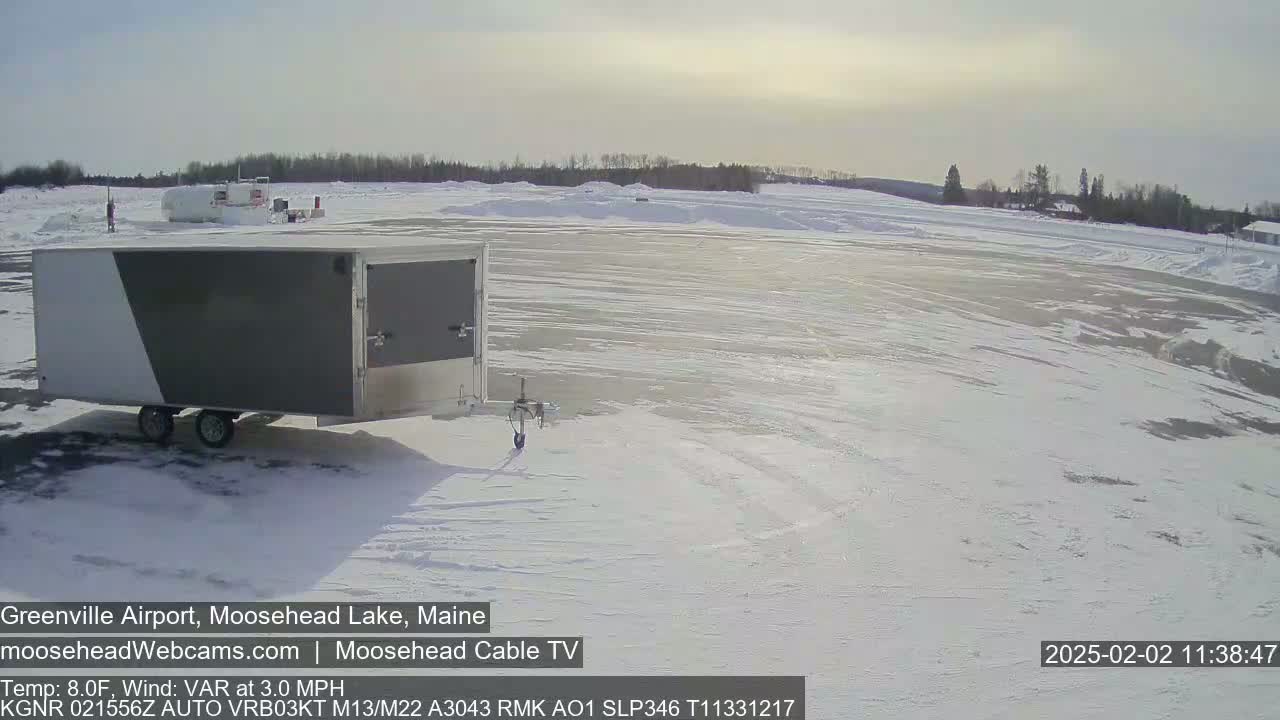 A gray and white enclosed trailer sits in a snow-covered airport parking lot under a mostly sunny sky.