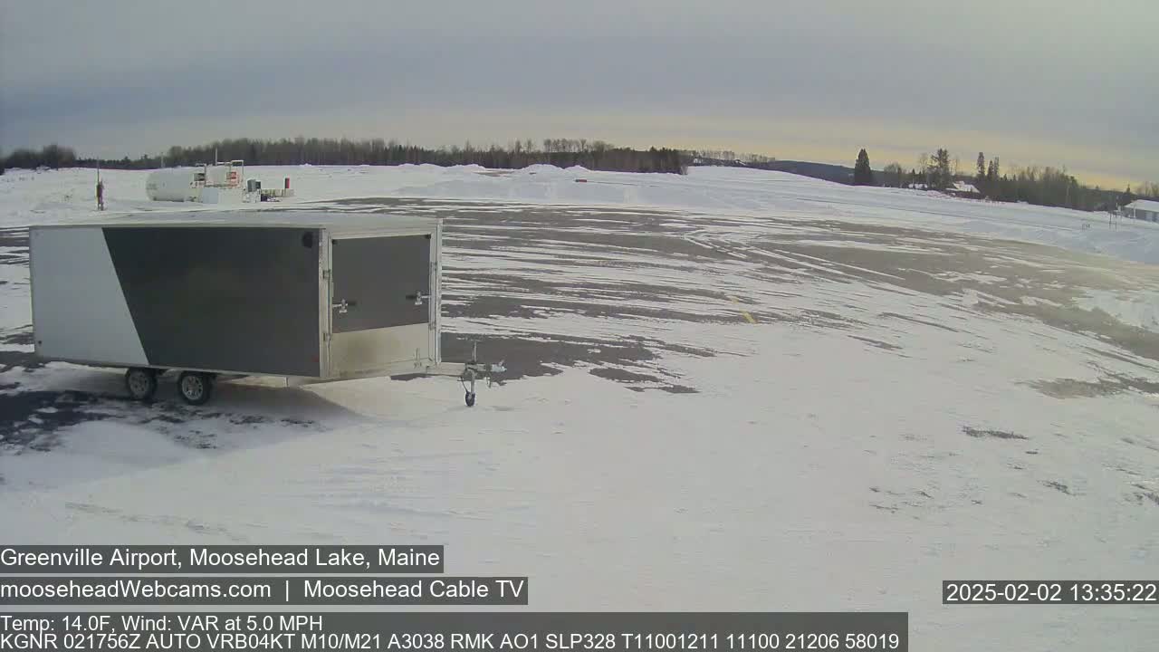 A gray and white enclosed trailer sits in a snow-covered airport tarmac under a cloudy sky.