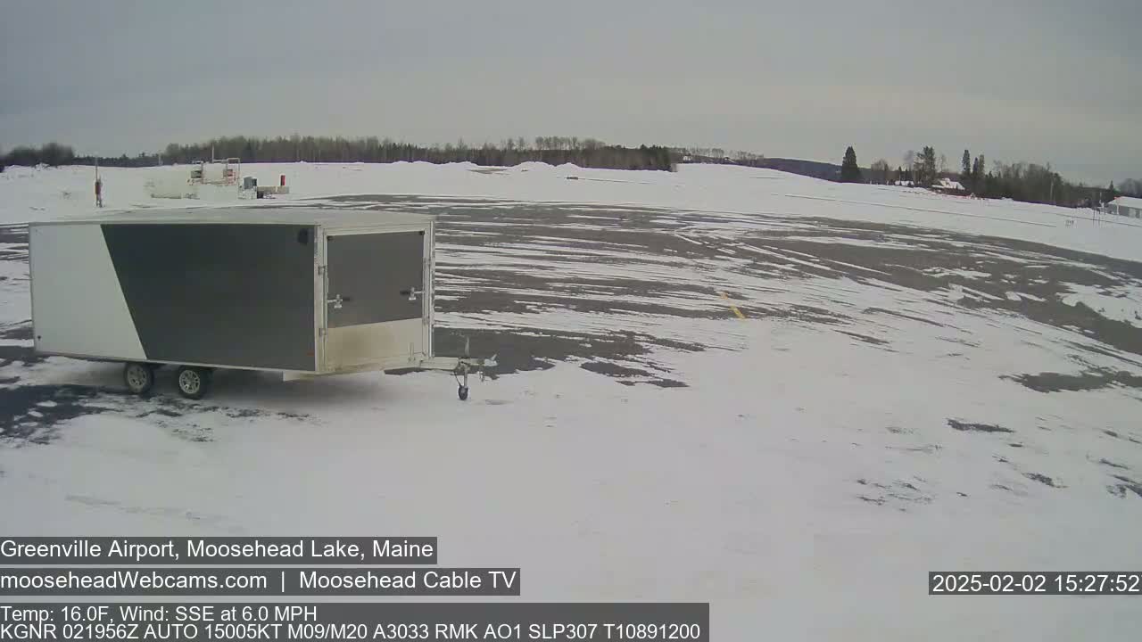 A gray and white enclosed trailer sits in a snow-covered airport parking lot under a cloudy sky.