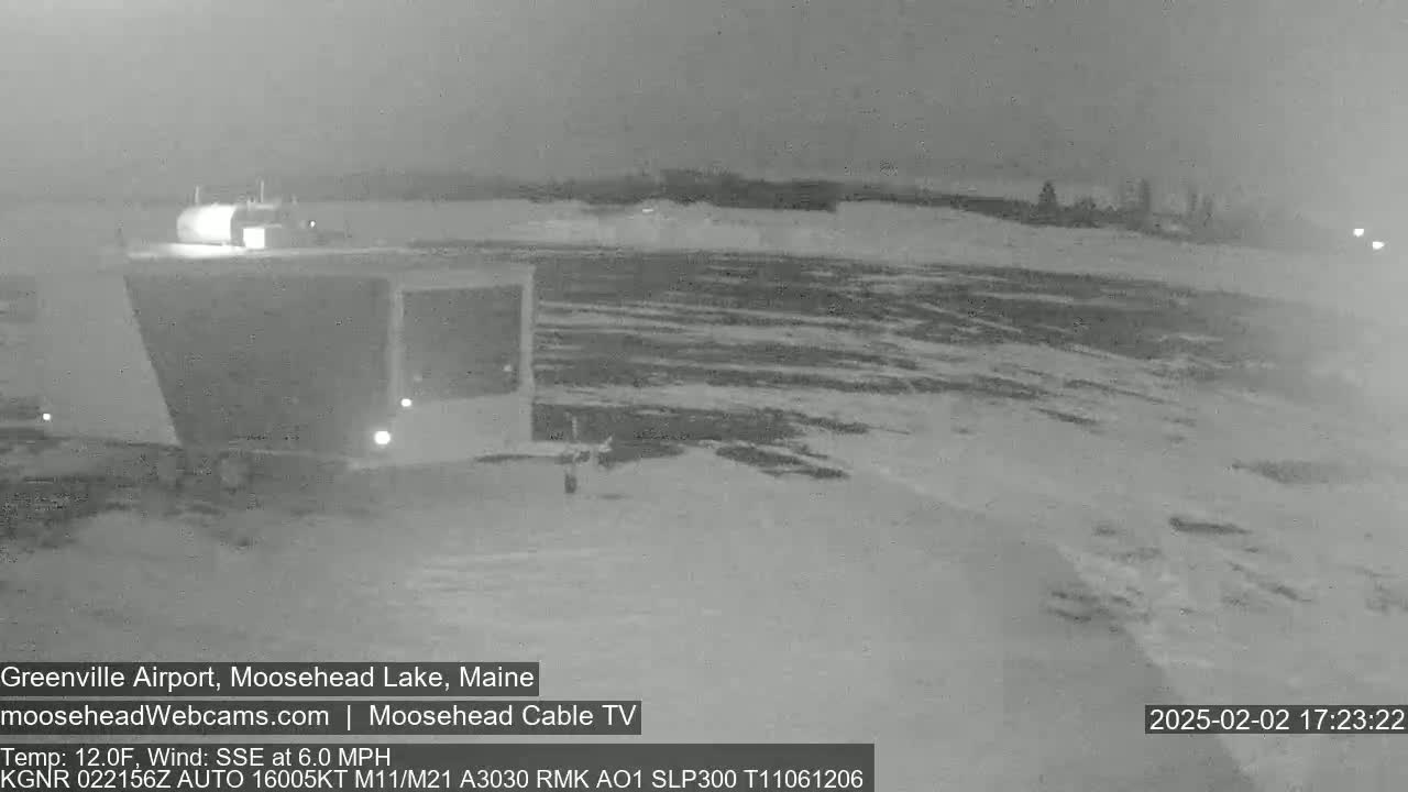 A nighttime, grayscale image shows a snow-covered airfield with a building and a vehicle under a dark, overcast sky.