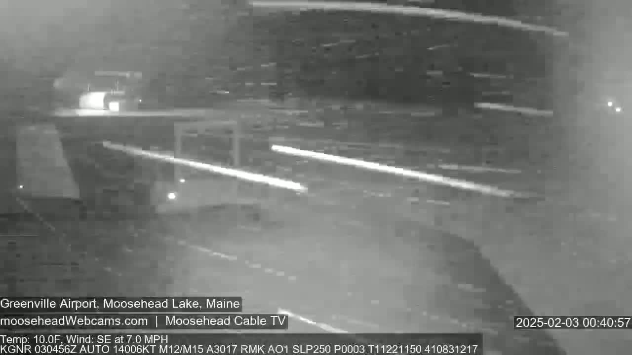 A nighttime, snow-covered road is shown with vehicle headlights visible in the distance, under windy conditions.