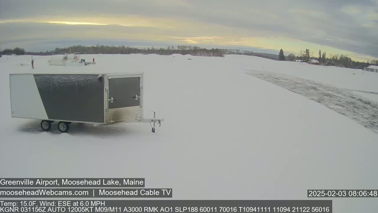 A gray and white enclosed trailer sits in a snow-covered airfield under an overcast sky.