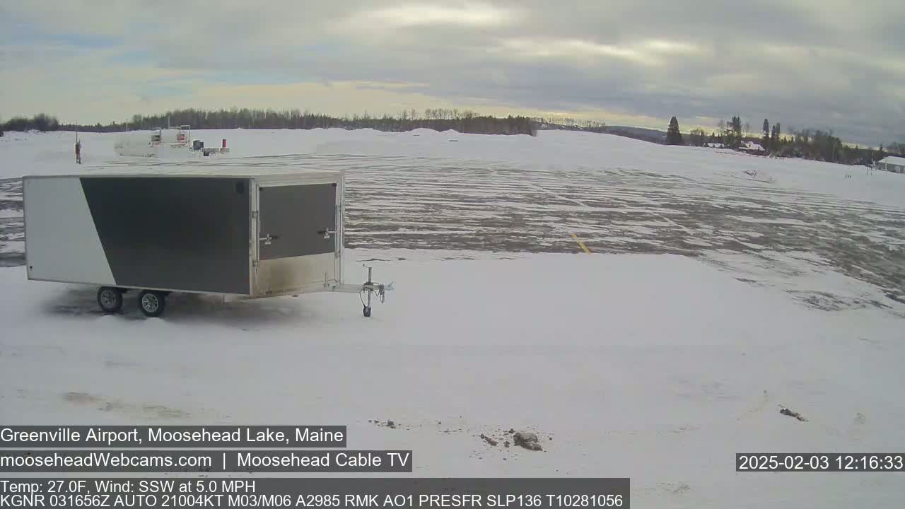 A gray and white enclosed trailer sits in a snow-covered airfield under a cloudy sky.