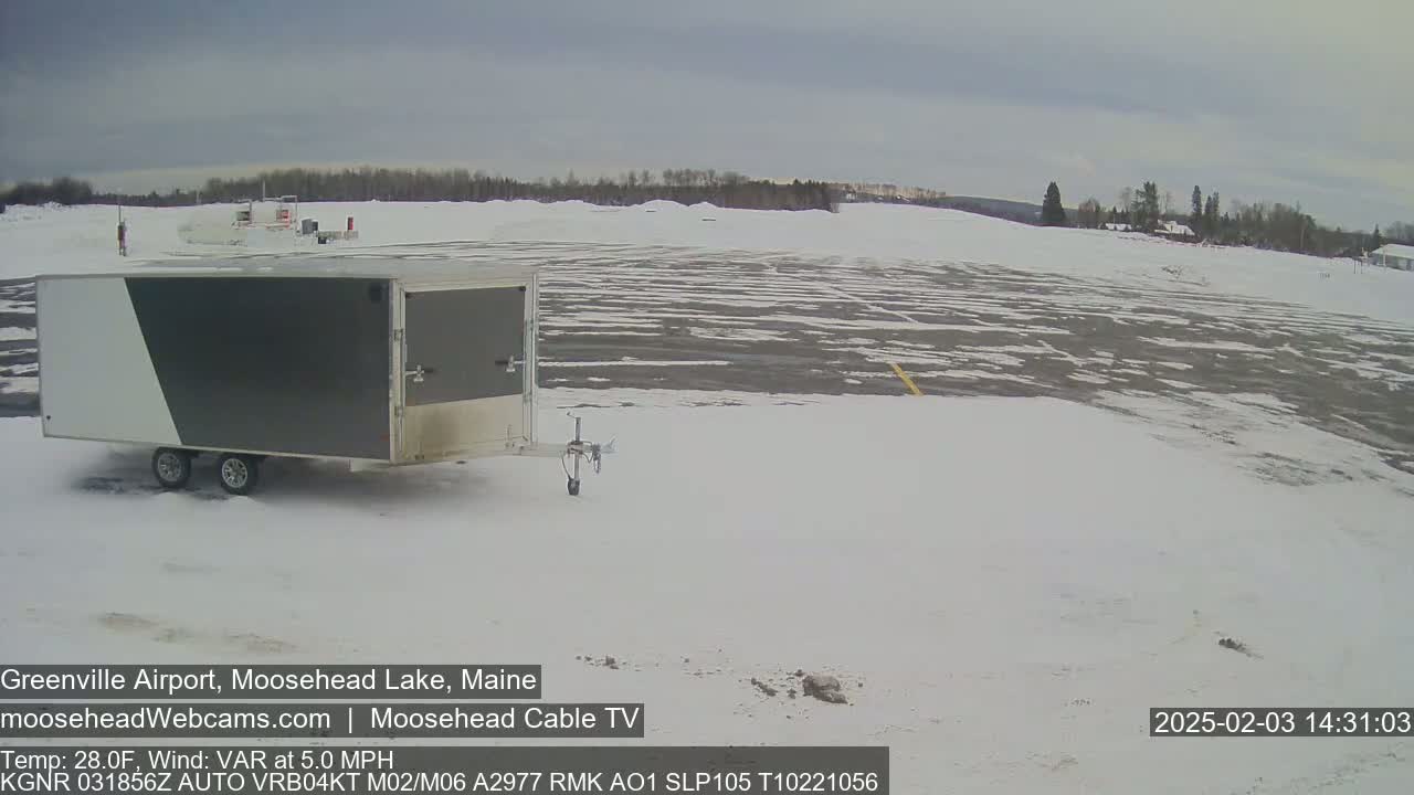 A gray and white enclosed trailer sits in a snow-covered airport parking lot under an overcast sky.