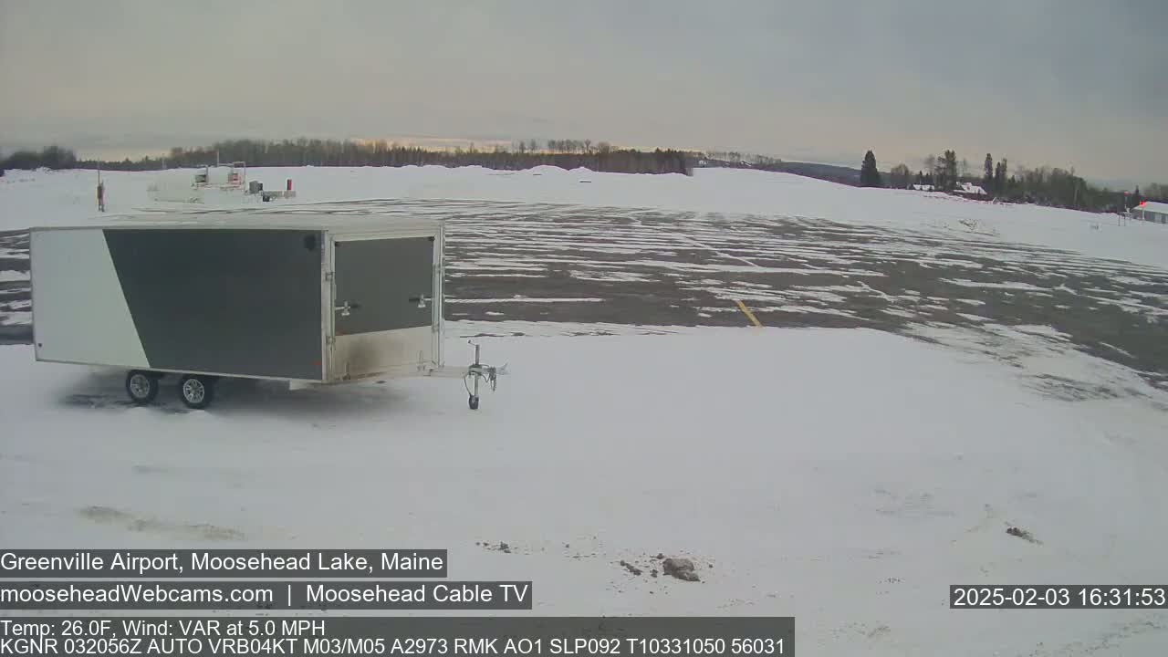 A gray and white enclosed trailer sits in a snow-covered airport parking lot on a partly cloudy day with a light wind.