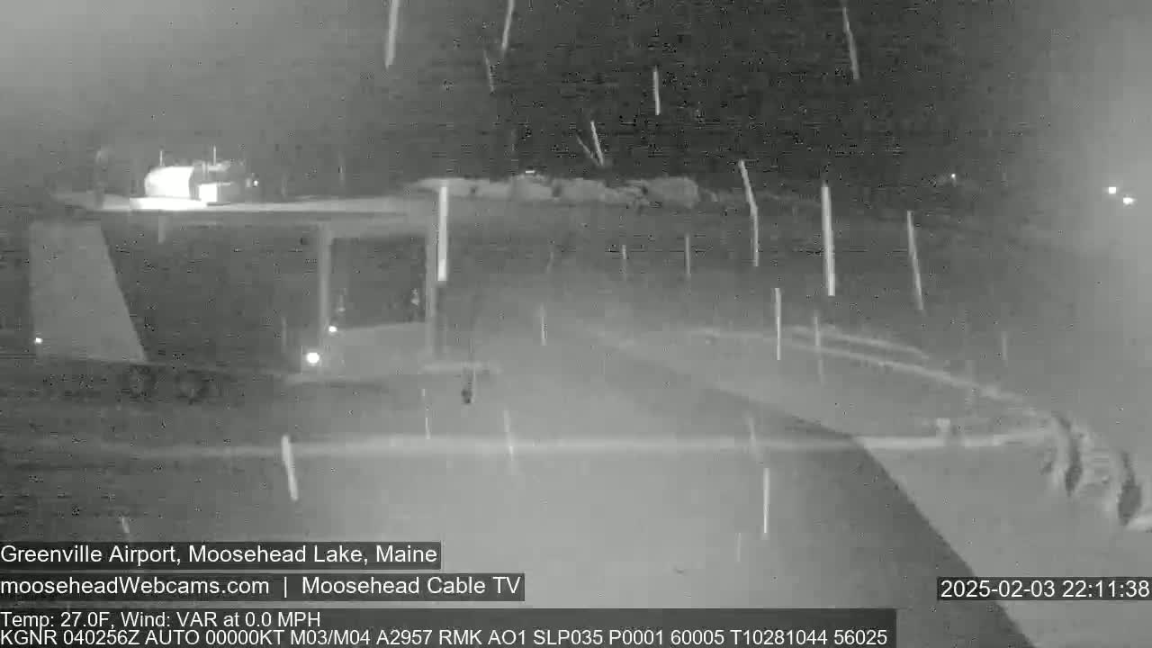 A nighttime, snow-filled scene shows a dark-colored vehicle partially visible in the upper left corner, with the falling snow obscuring much of the view.