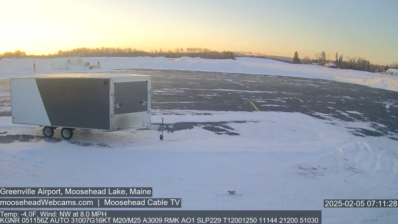 A gray and white enclosed trailer sits in a snow-covered airport parking lot at sunrise, with a calm northwest wind.